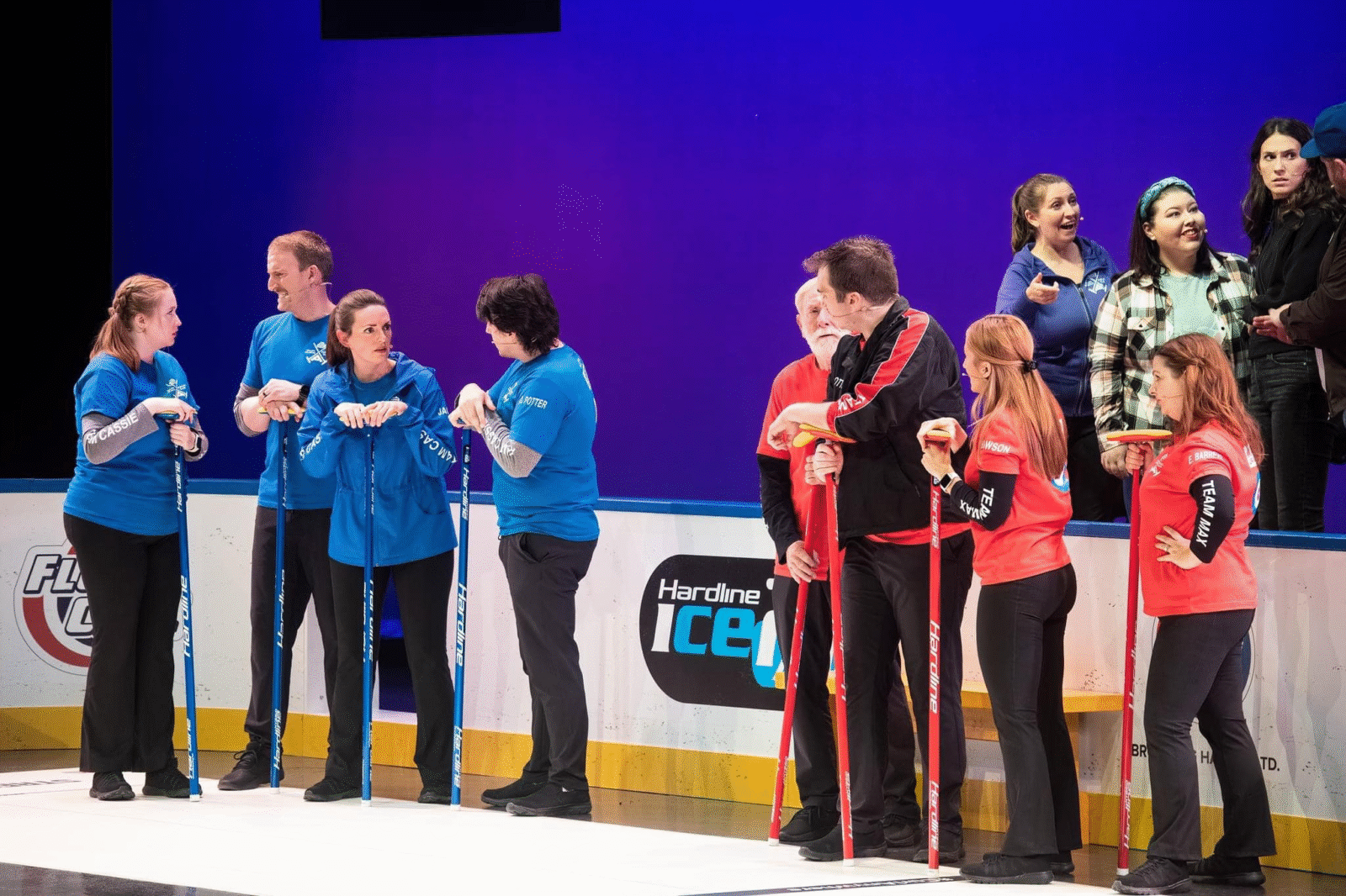 Production photo from 'Second Shot' the musical: two curling teams - one in red and one in blue - talk smack about each other while fans look on in the stands.